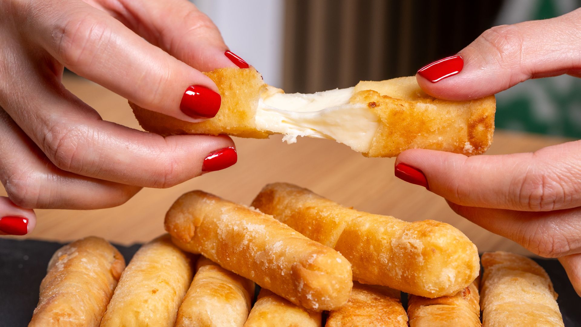 Close-up of a woman's hands with red nails breaking cheese sticks on a slate board.