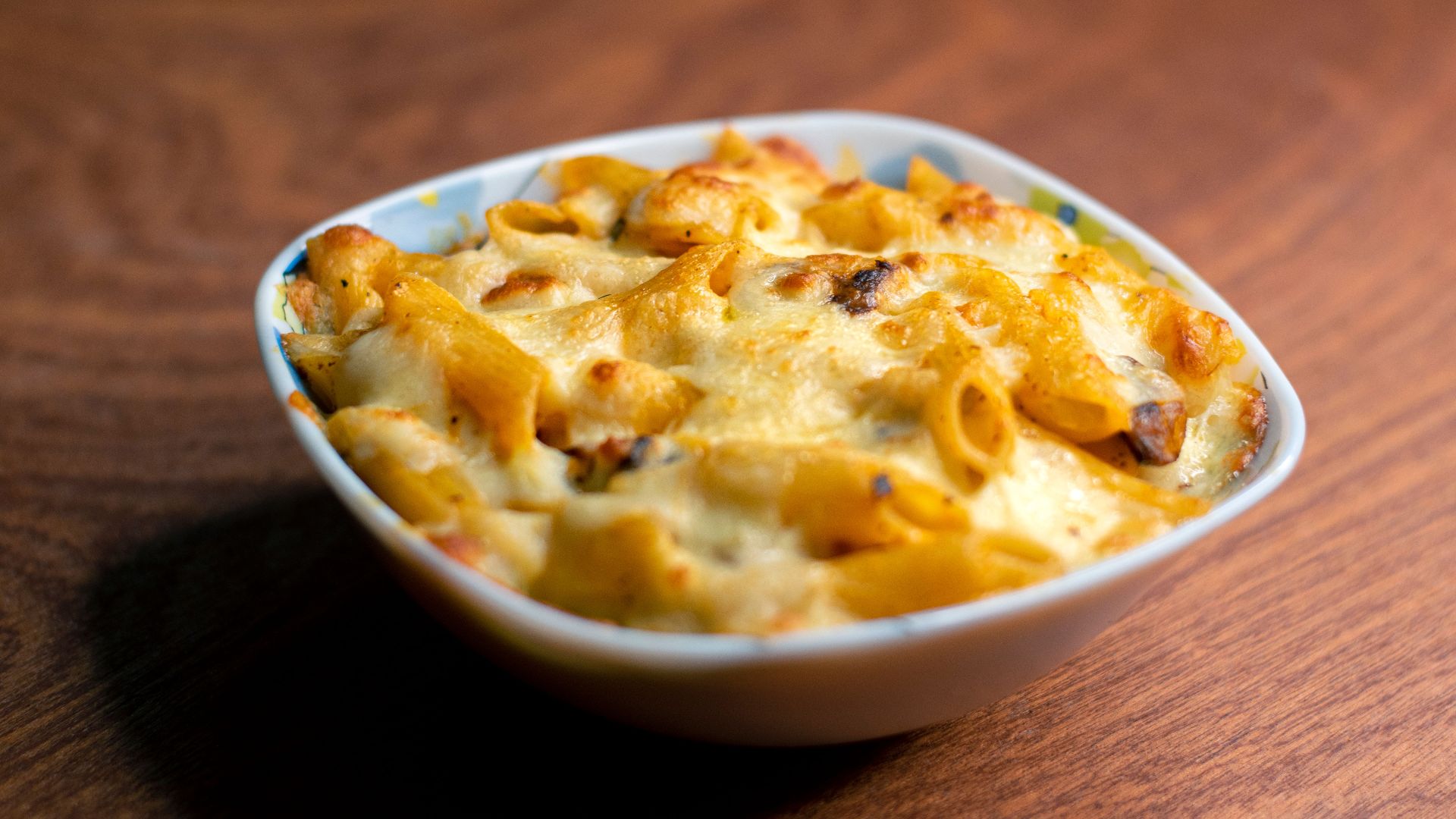 A close-up of cheesy baked macaroni in a white bowl on a wooden table.