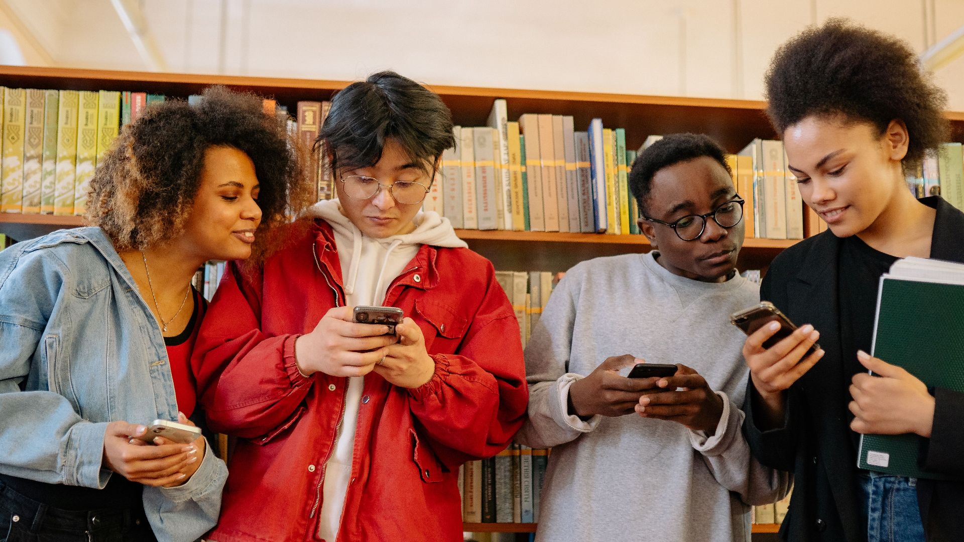 A group of diverse young adults using smartphones while interacting in a library setting.