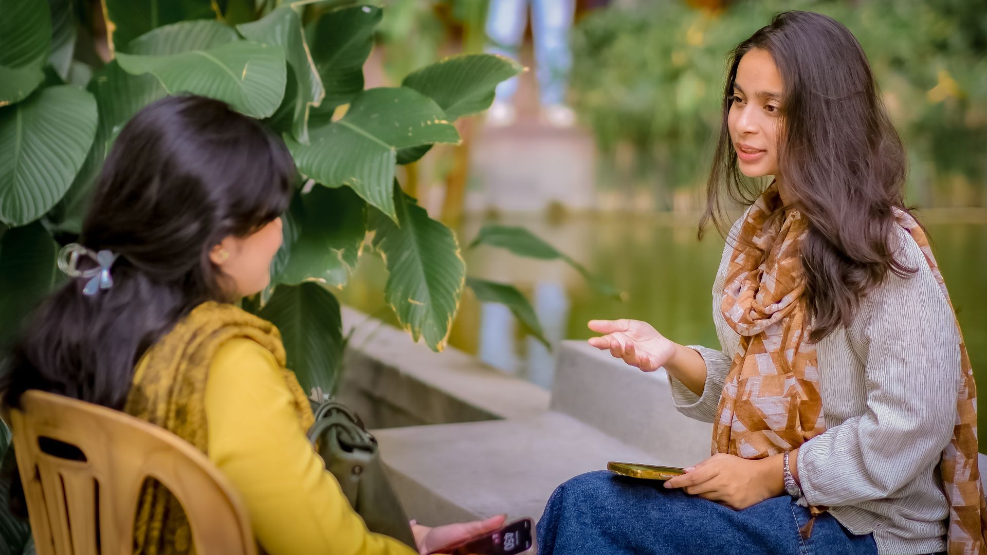 Two women sitting near lush plants having a conversation outdoors. Relaxed and candid atmosphere.