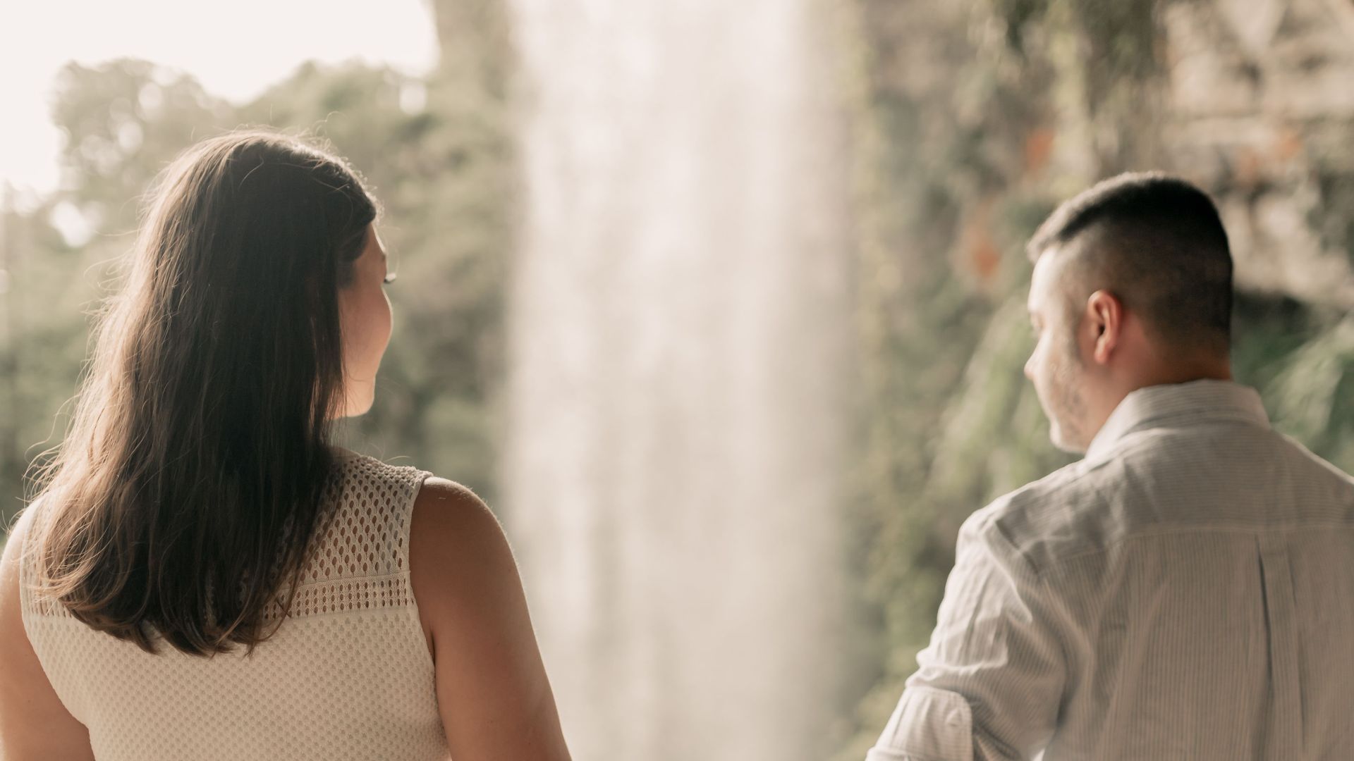 Romantic couple holding hands near a waterfall amidst lush greenery.