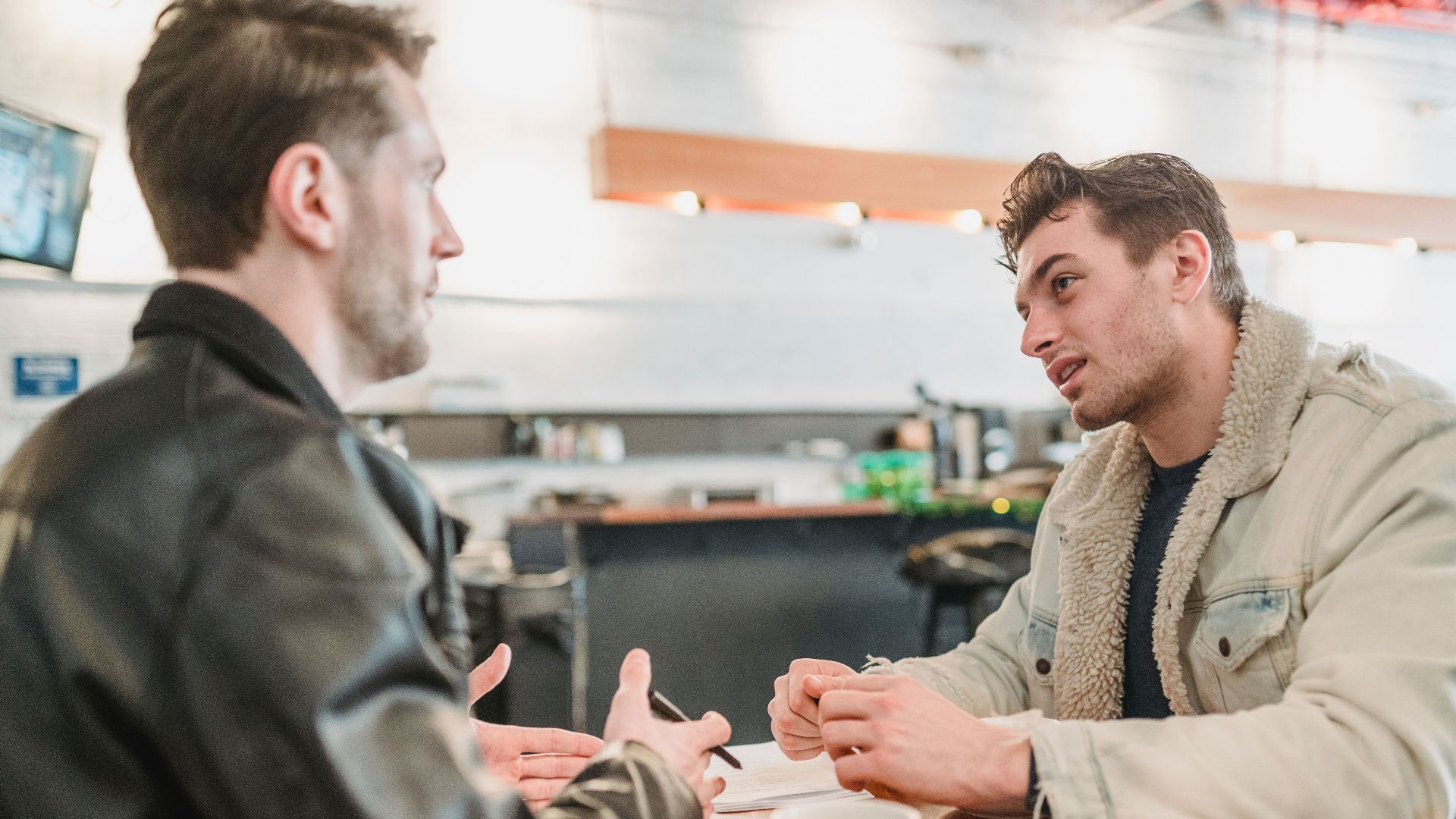 Side view of crop young man talking about business project at table with papers and looking at partner on blurred background