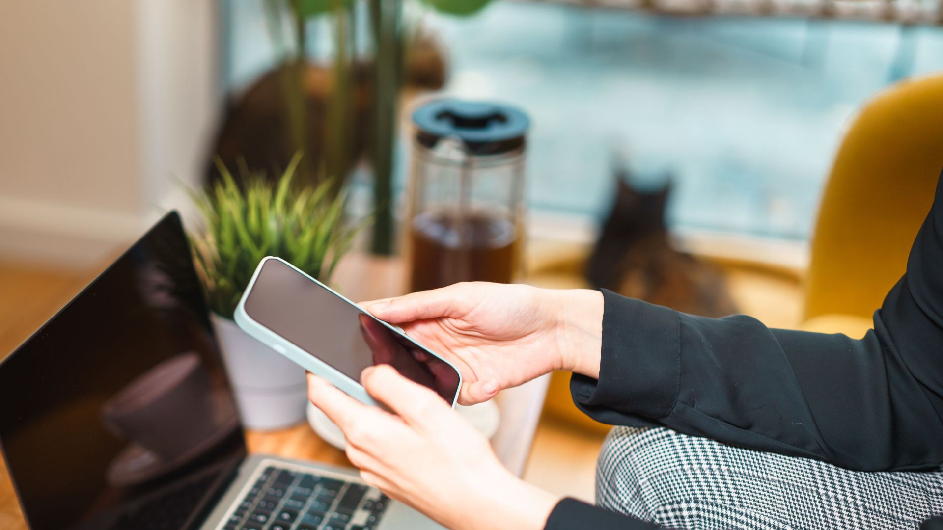 Woman working remotely from home, using a smartphone and laptop at a cozy desk with a plant and cat in background.
