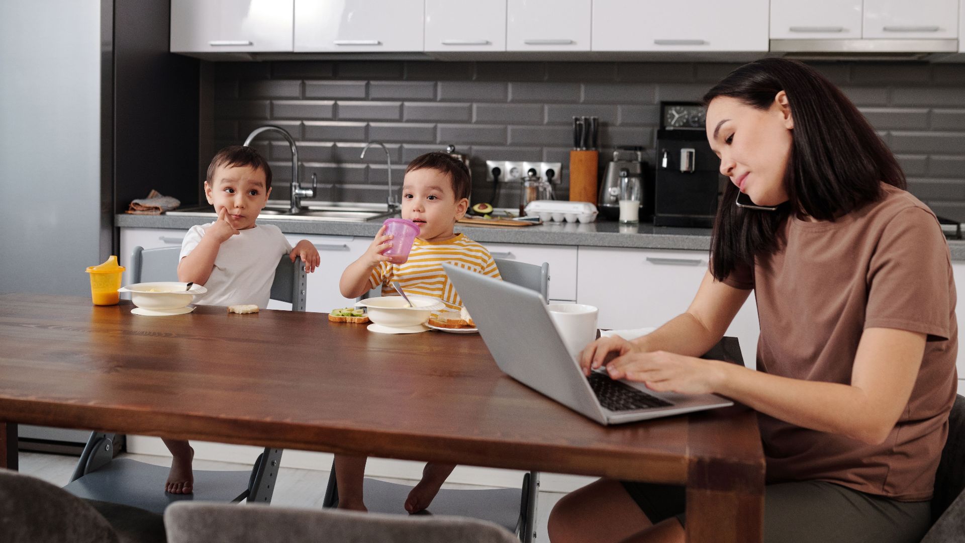 A mother multitasking on a laptop while her two young sons enjoy breakfast in the kitchen.
