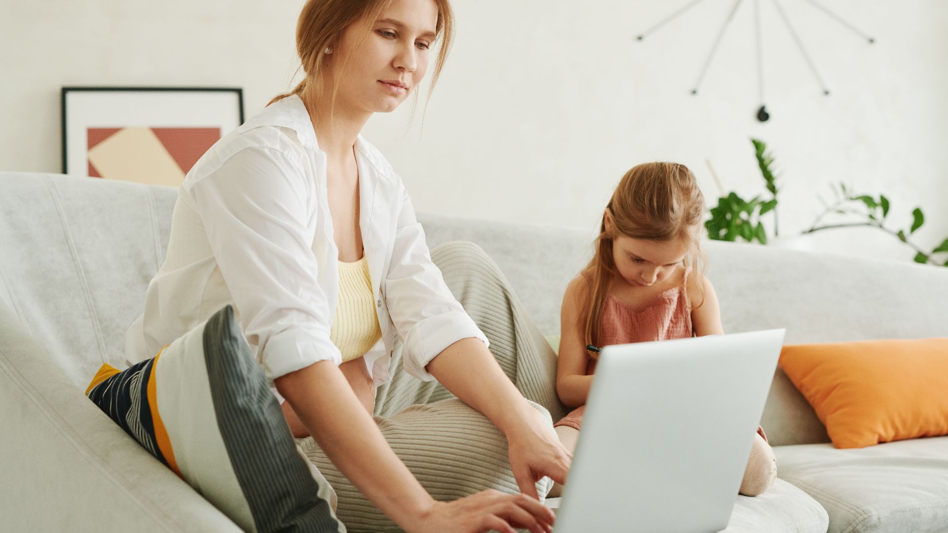 A mother and daughter use a laptop together on a cozy sofa indoors, multitasking family life.