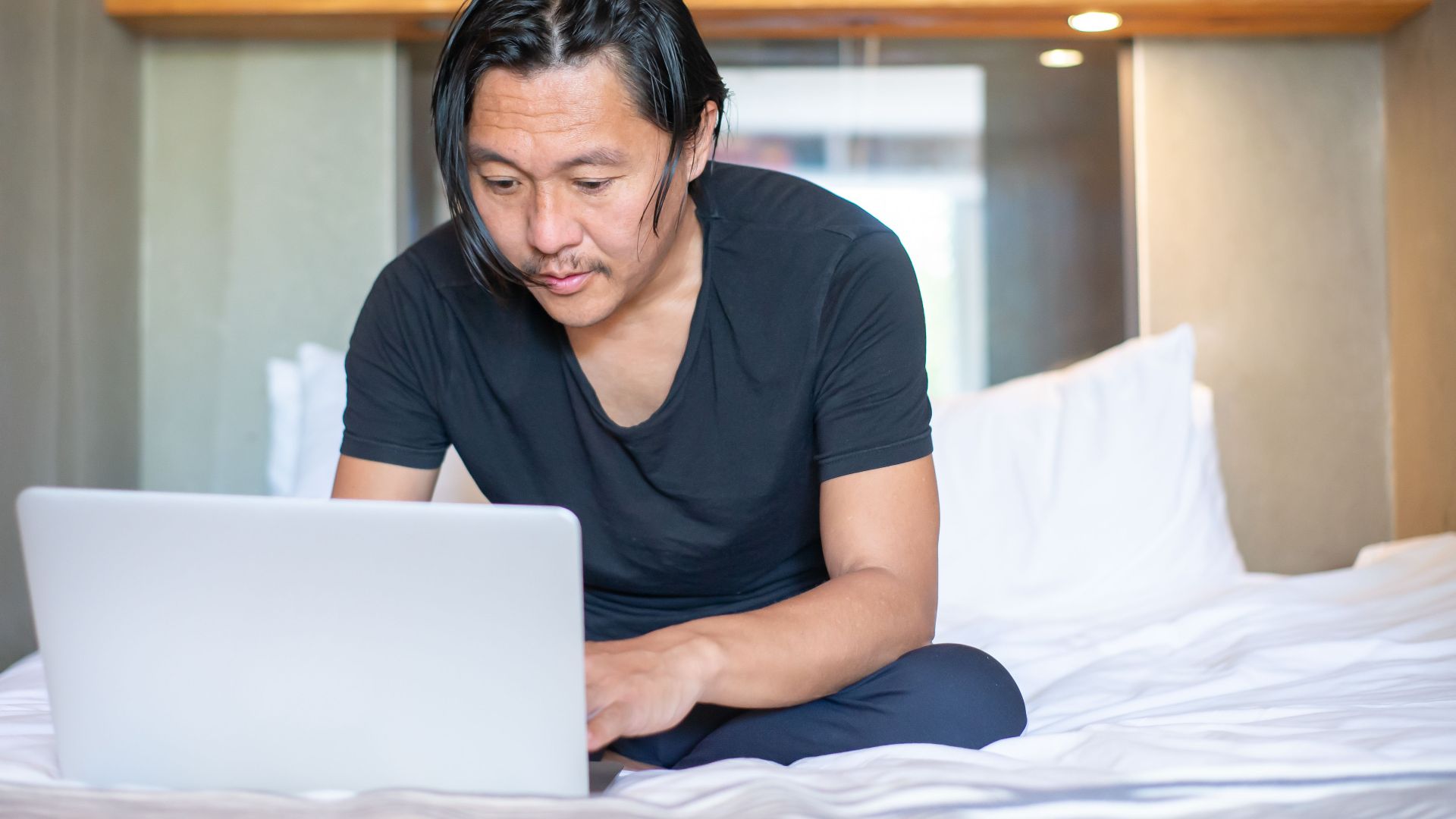 Asian man working remotely on a laptop in a cozy bedroom setting, exemplifying modern work-from-home lifestyle.