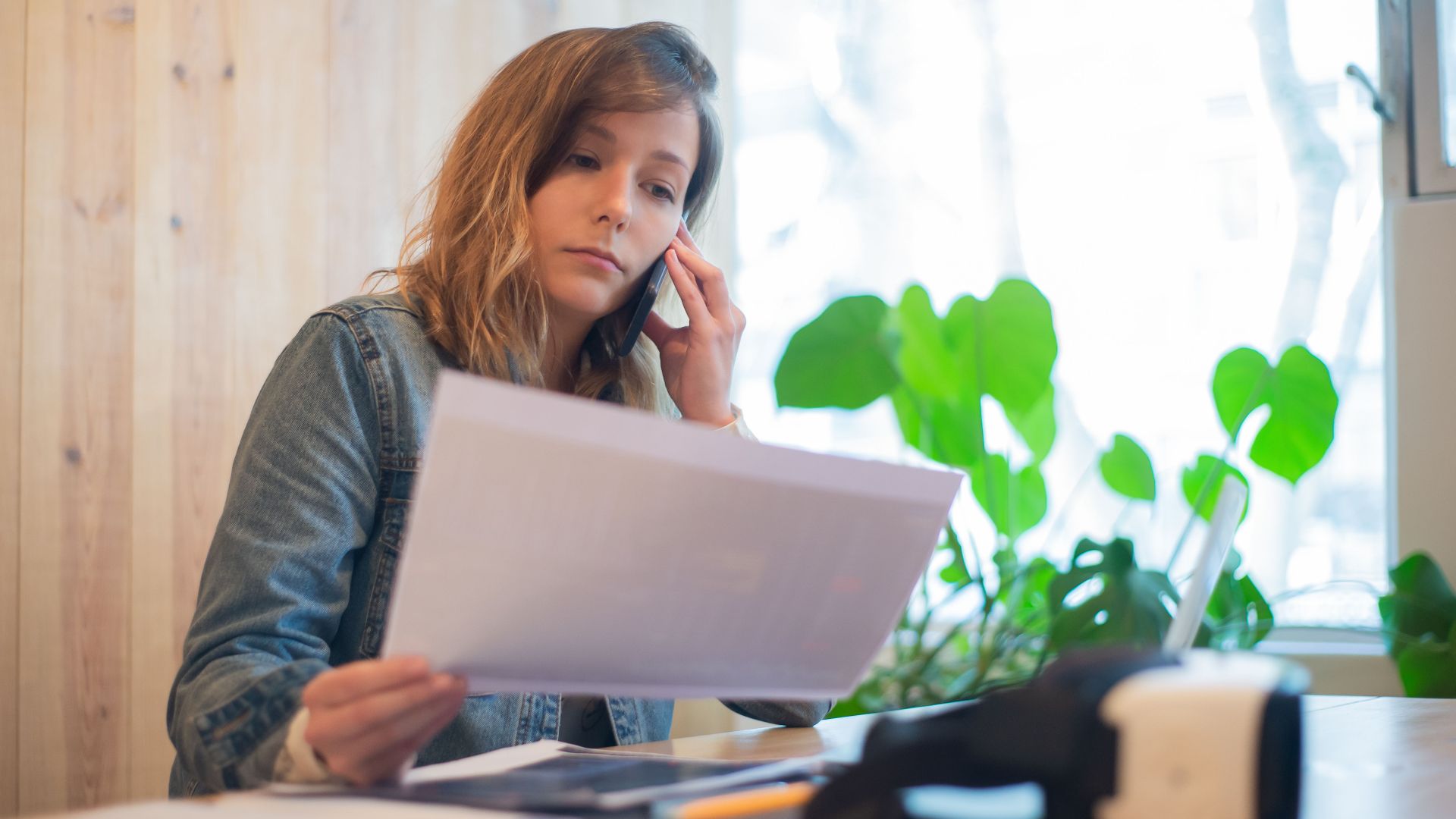 Businesswoman reviewing paperwork while on a call at a modern office workspace.