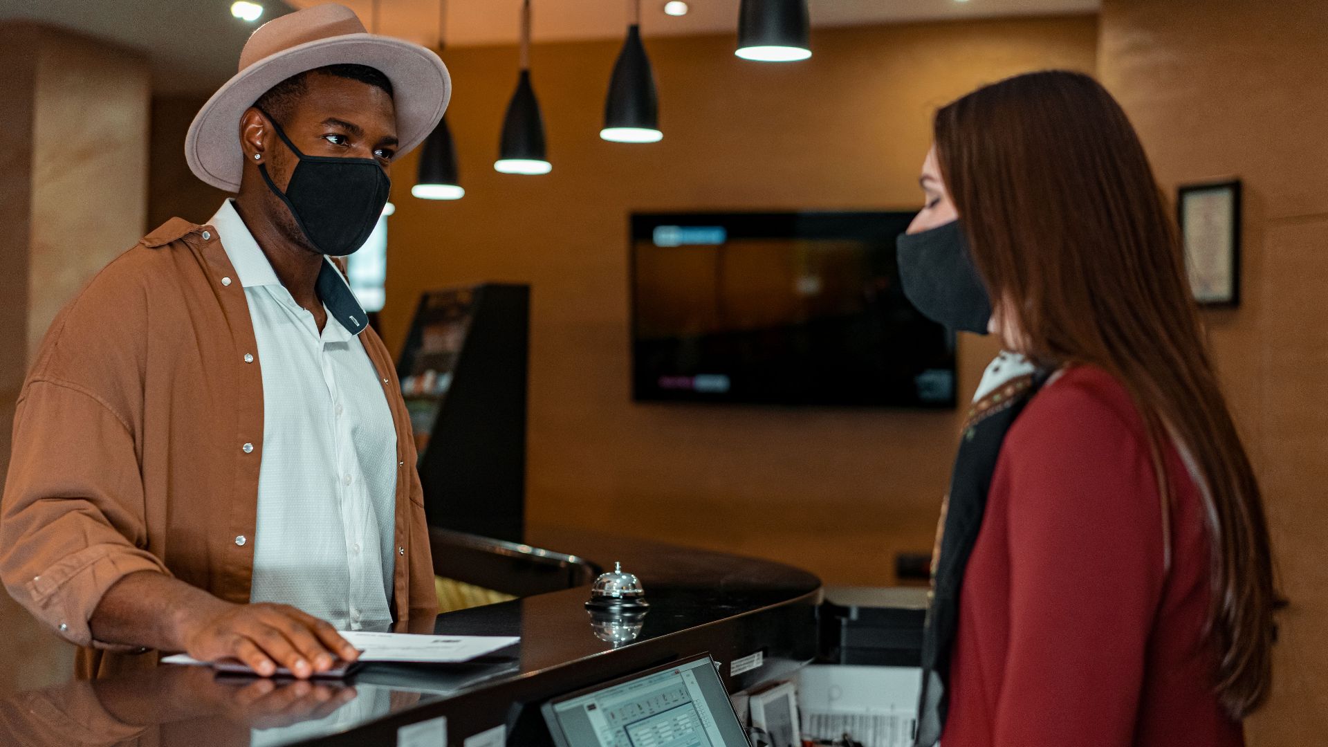 A man and woman in face masks checking in at a hotel reception, emphasizing safety protocols.