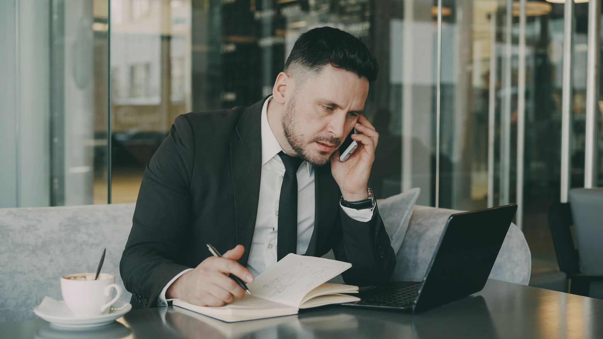 Businessman talks on phone while writing notes in a modern cafe.