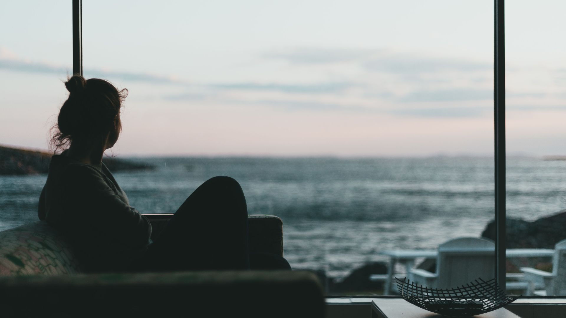A woman sits indoors, viewing the ocean through a glass window at sunset, creating a serene silhouette.