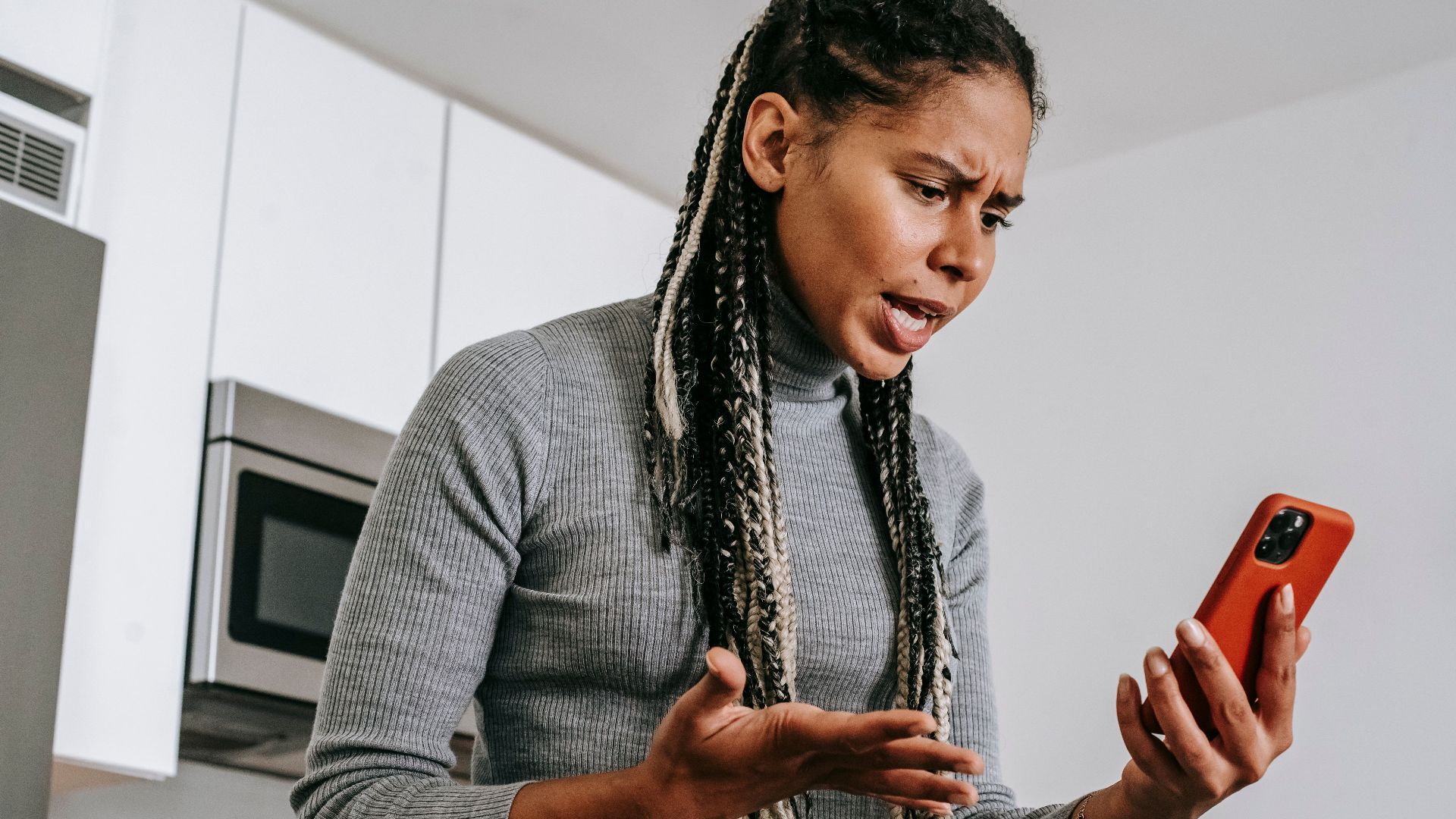 From below of angered black female with Afro braids in casual outfit gesticulating and resenting while having video call on smartphone in modern light kitchen