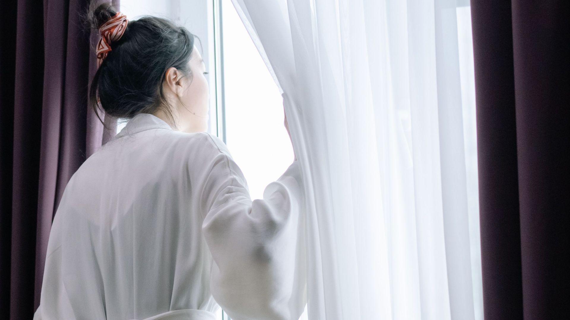 A woman standing in a bathrobe looks out the window, creating a serene indoor moment.