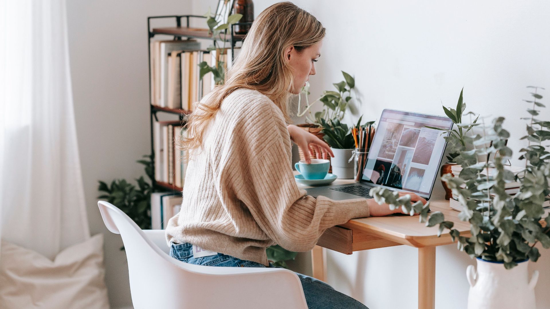Side view of attentive female remote employee with cup of tea watching photo gallery on netbook screen at desk in house