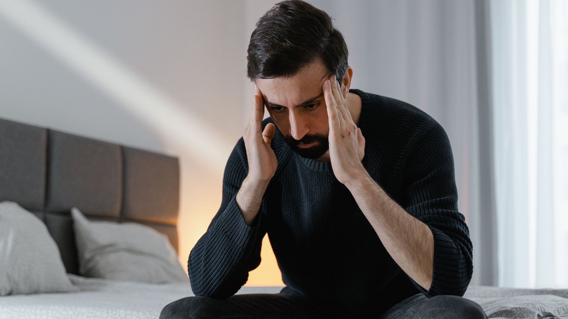 A man in a black sweater sits on a bed, expressing frustration in a bedroom.