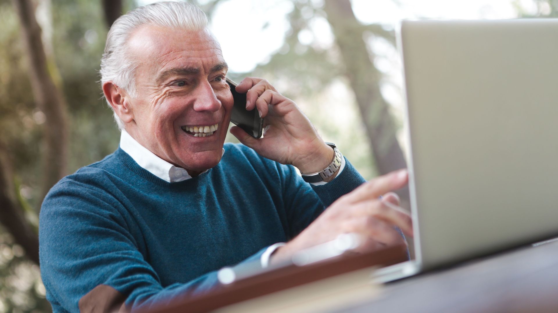Cheerful senior man working remotely, talking on phone and using laptop outdoors.