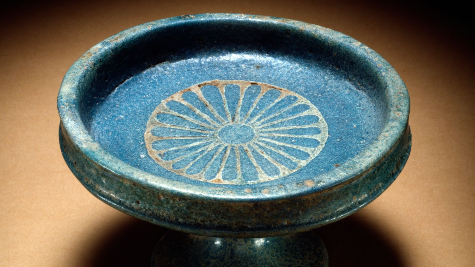 A daisy decorates the interior of a tazza, or footed dish.  A point on the bottom of the saucer fits into a corresponding depression in the stand, securing the two together.  Patterns cut into the still moist composition were filled with a different color