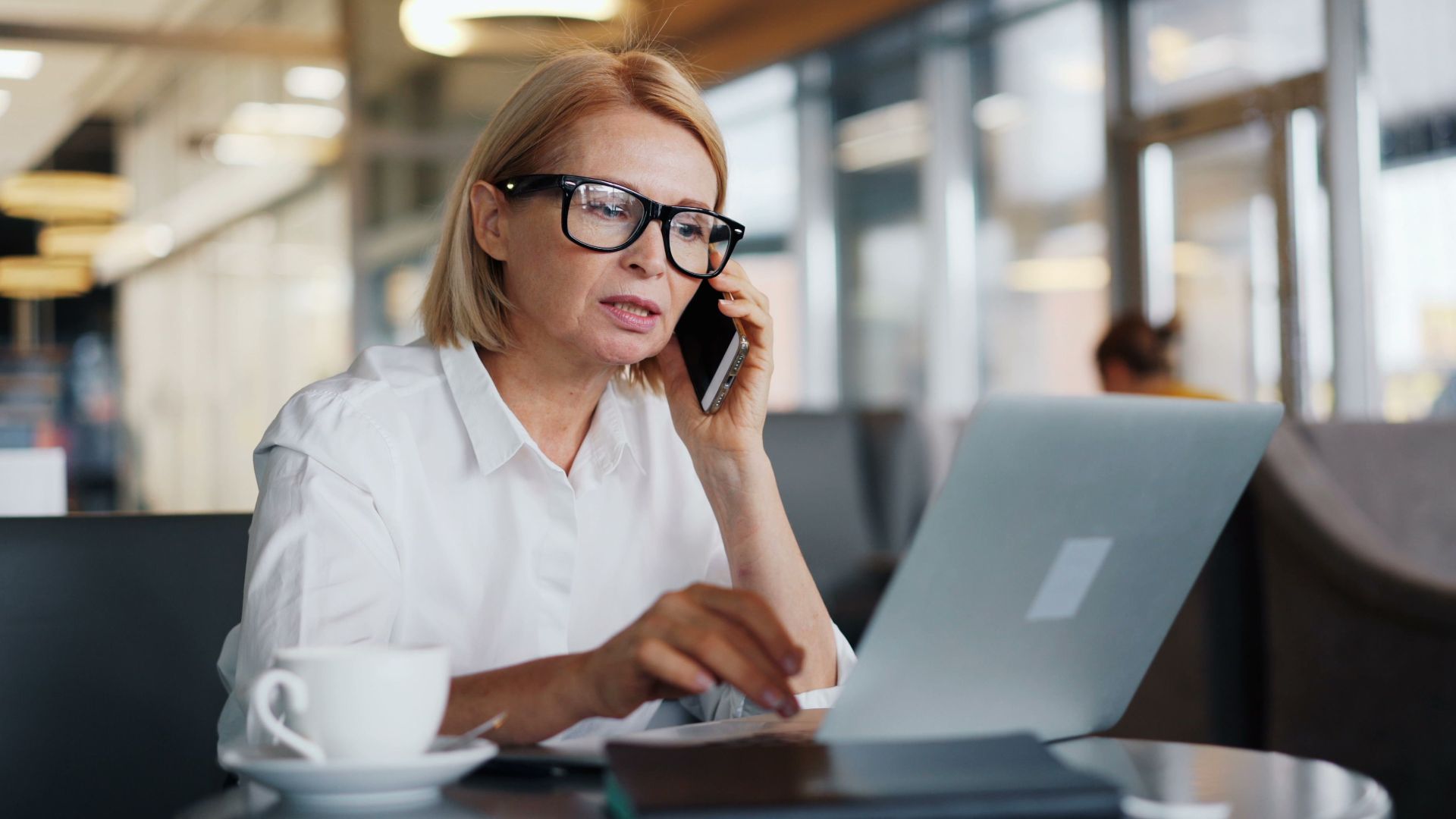 Mature woman in a café using a laptop and smartphone for business communication.