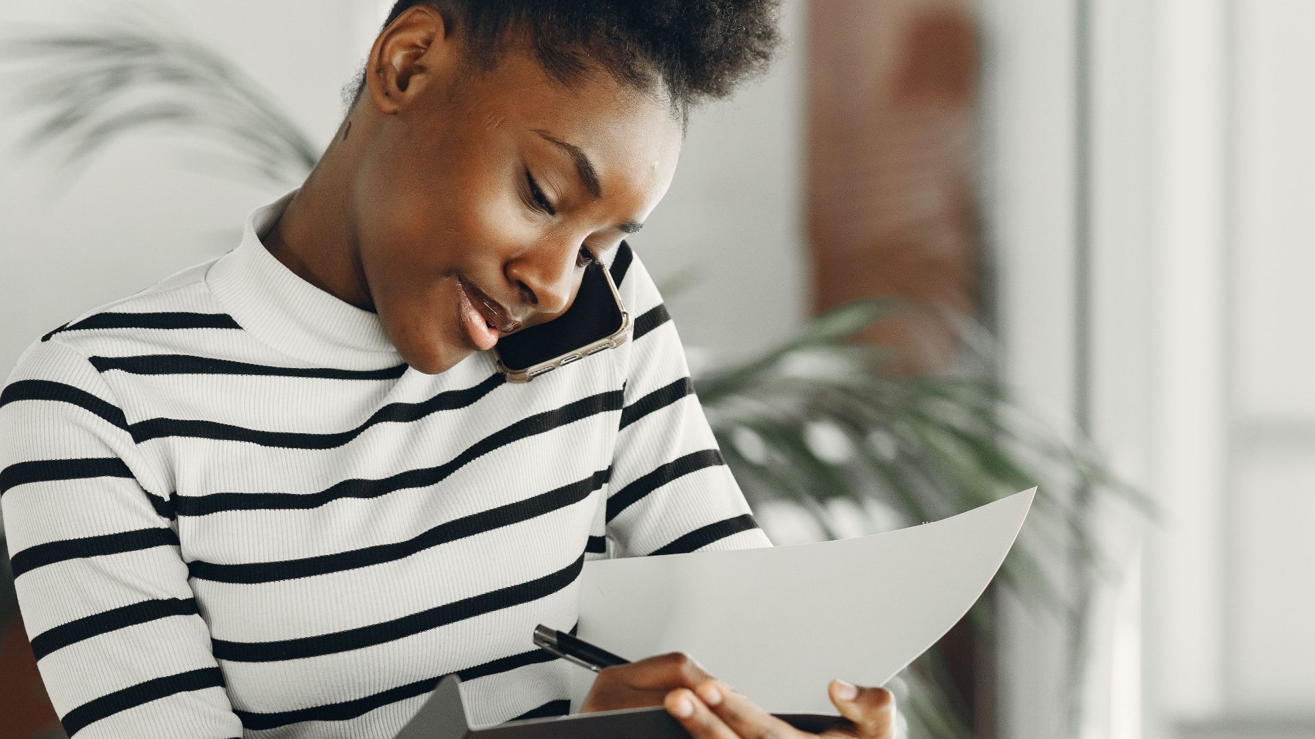 Young African woman multitasking with a phone call and taking notes indoors, wearing stripes.