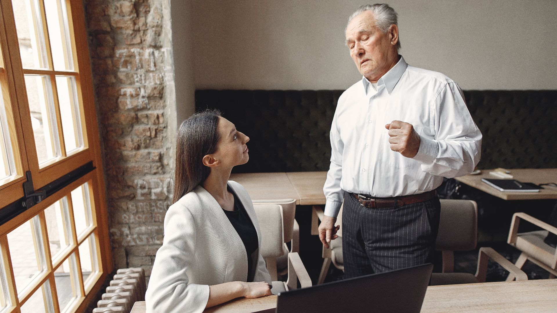 Aged businessman in formal wear talking with well dressed female employee with laptop and cup of coffee about business issue in modern office