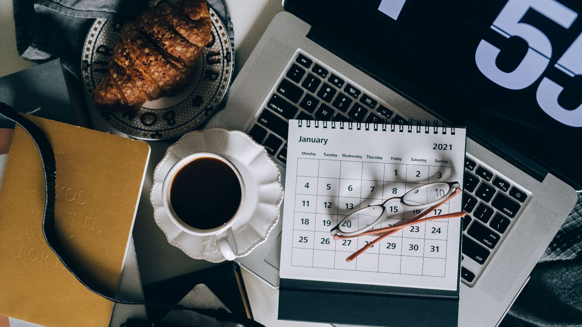 A serene workspace setup with coffee, croissant, laptop, and calendar, ideal for organizing plans.