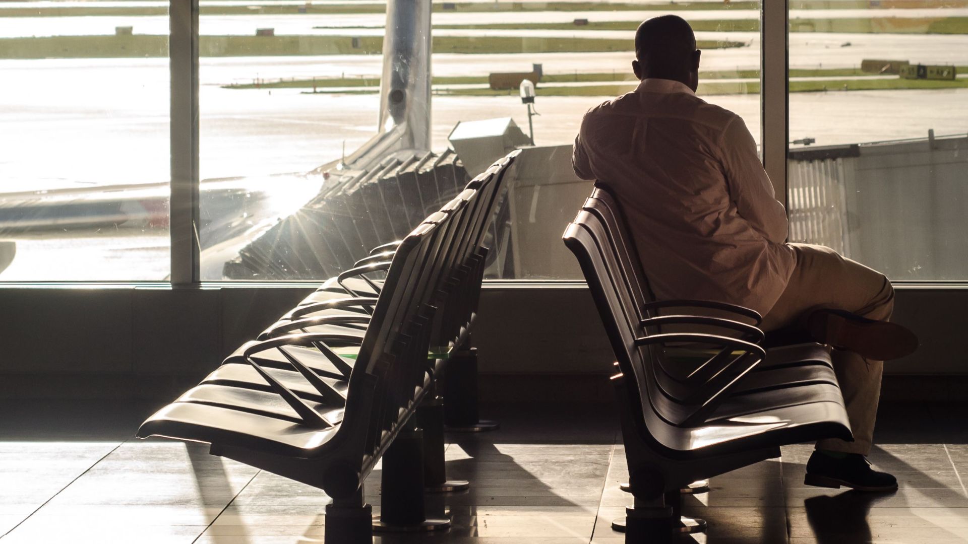 A man sits in an airport terminal, watching an airplane through the glass at sunset.
