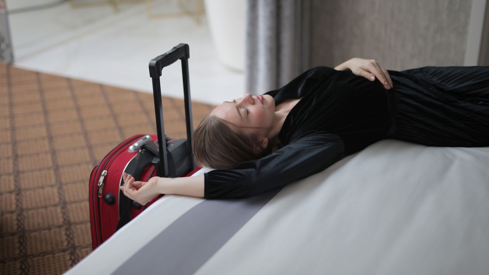 A woman resting on a hotel bed beside her suitcase, capturing travel fatigue.