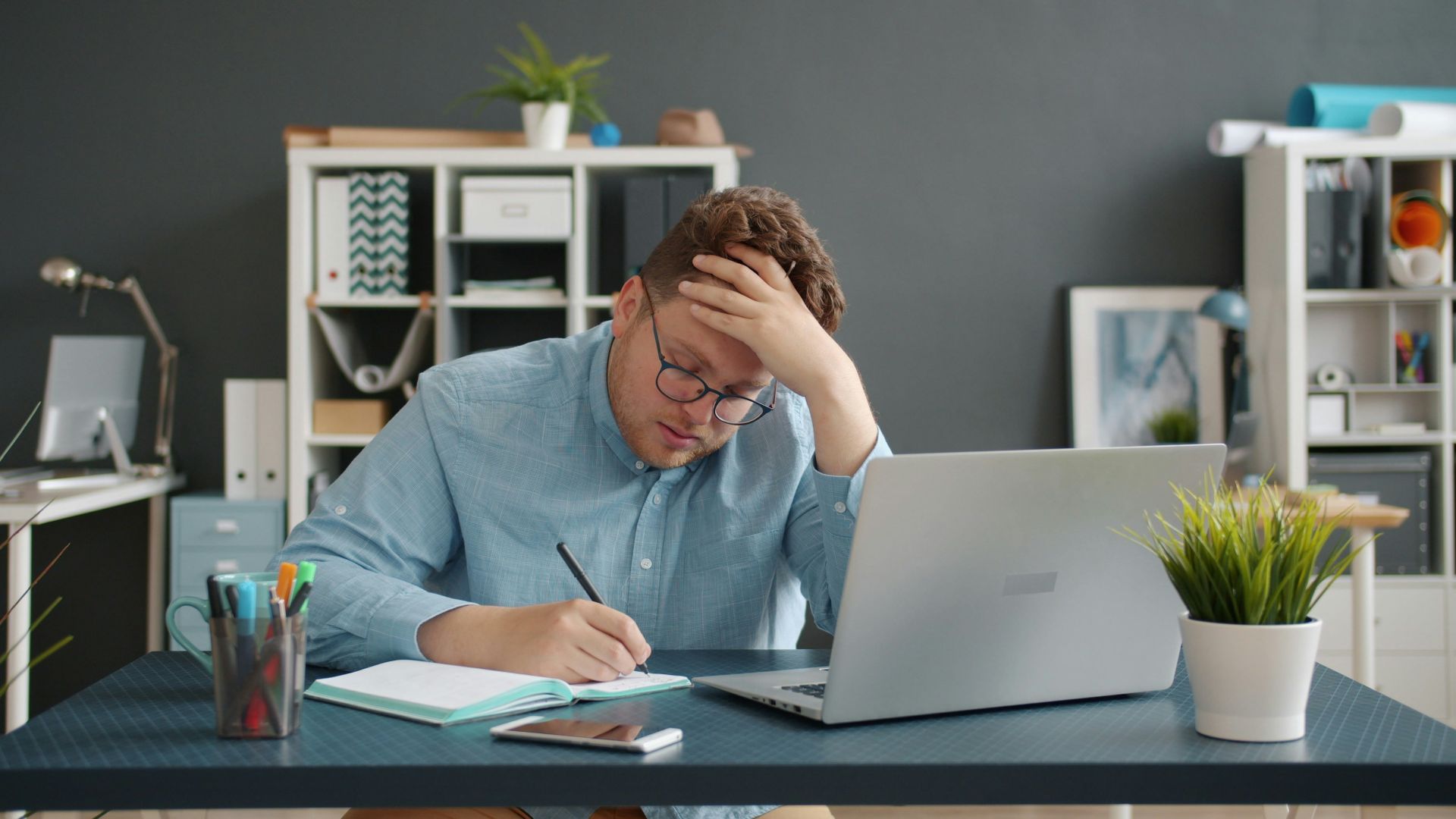 Man writing at desk with laptop, looking stressed