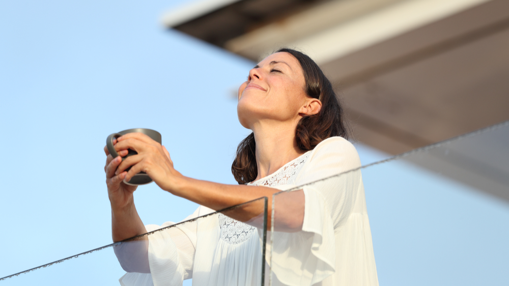 Satisfied adult woman with coffee cup breathing fresh air on a hotel balcony at summer