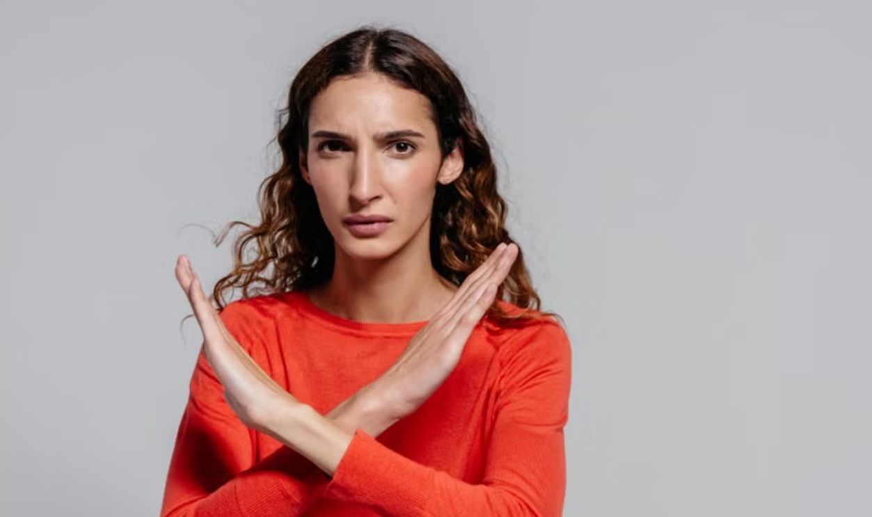 Woman with crossed arms making an X gesture against a gray background, indoor portrait.