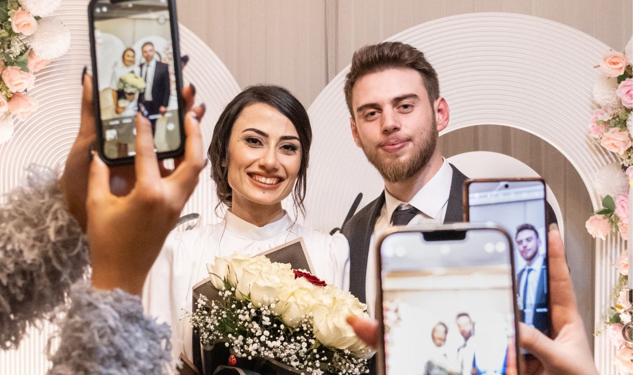 Joyful bride and groom pose with friends capturing memories on smartphones at their wedding celebration.