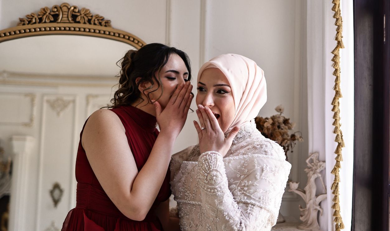 Two elegant women in formal attire whispering to each other, creating a sense of intrigue.