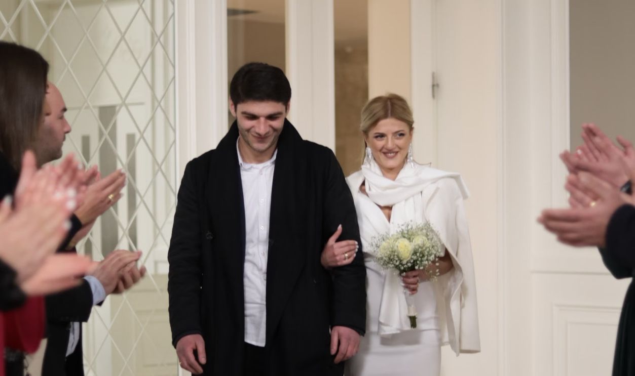 A newlywed couple smiling and walking arm-in-arm surrounded by applauding wedding guests.