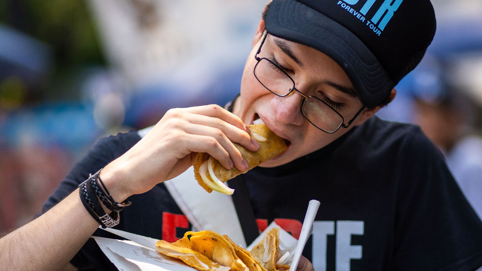 A young man wearing a cap and glasses enjoys street tacos in Mexico City.