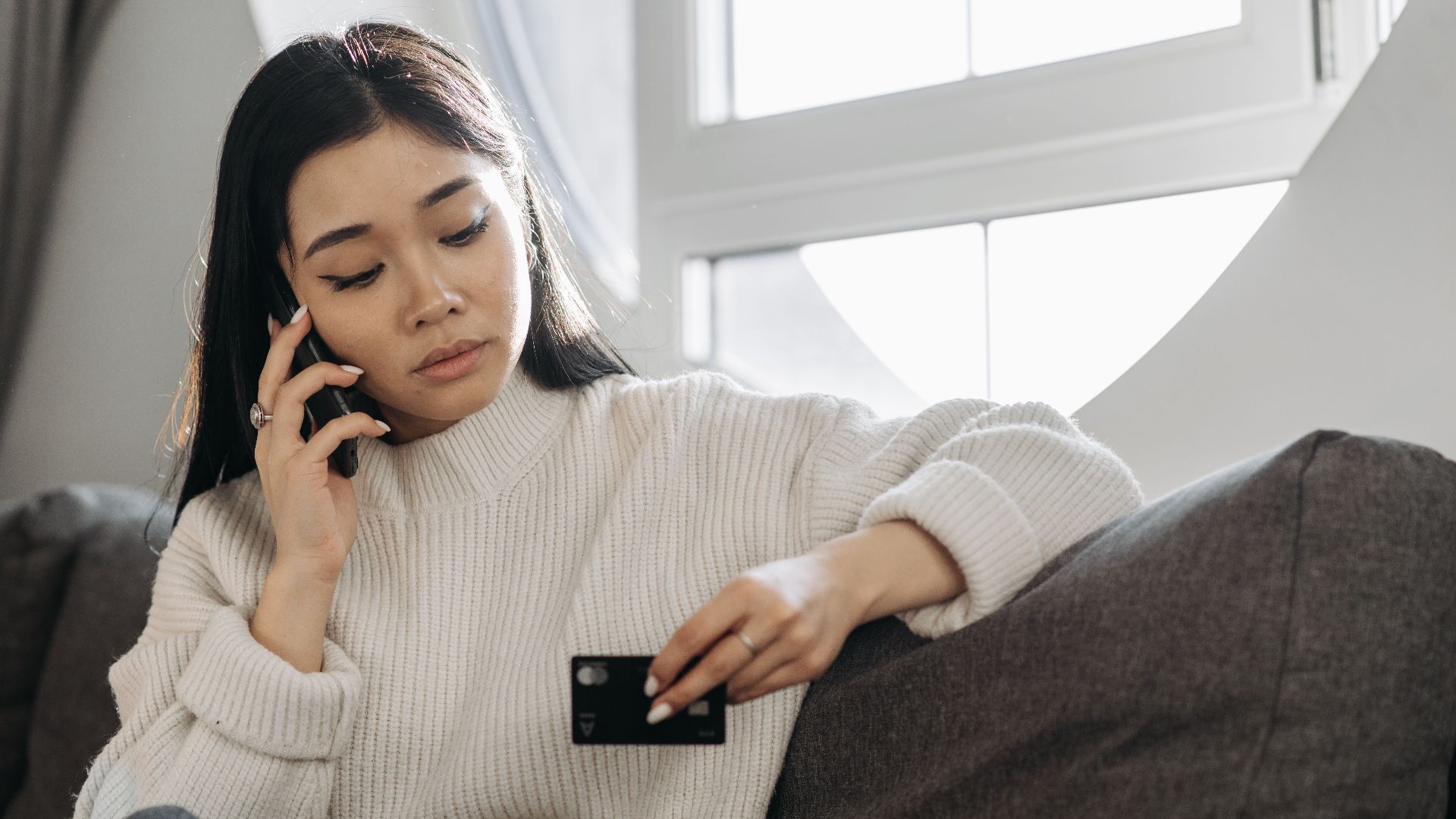 A woman sits on a sofa, multitasking with a phone and credit card, focusing on online shopping.