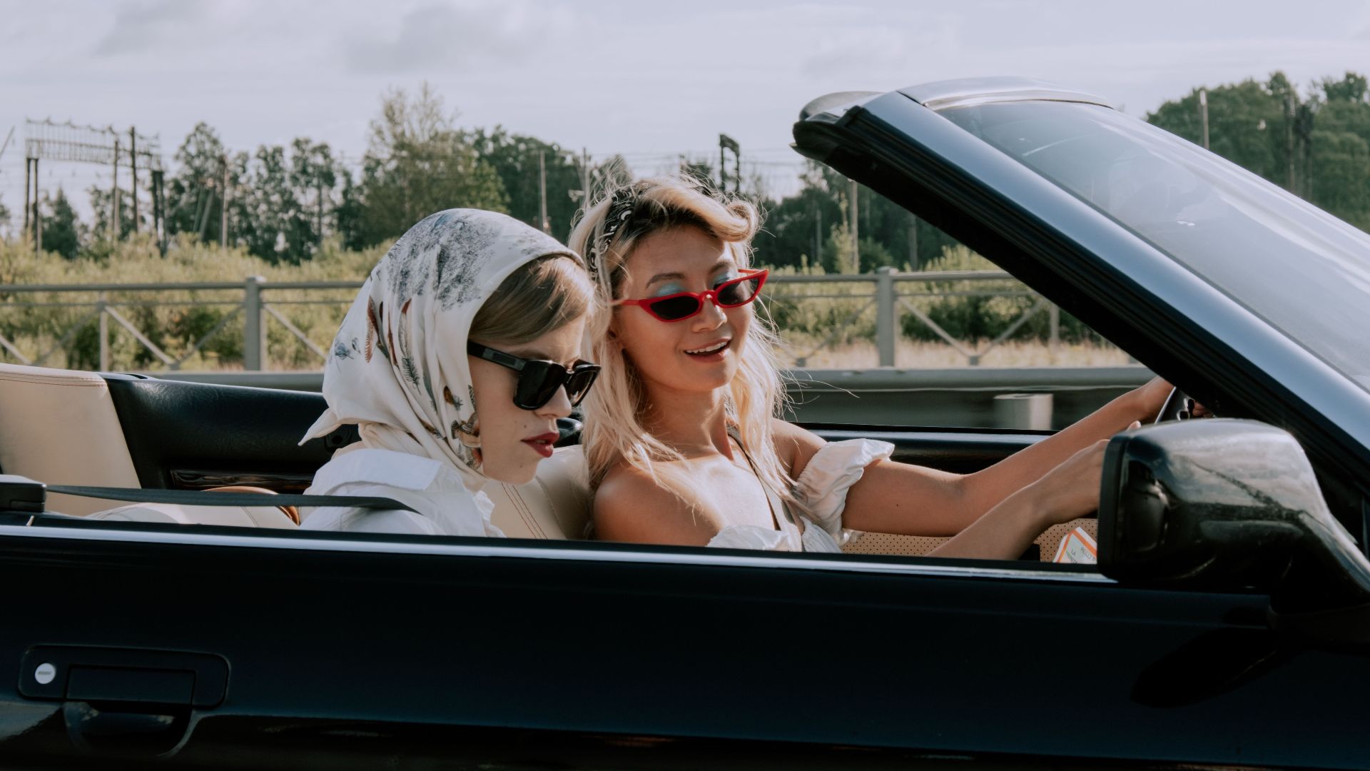 Two stylish women driving a convertible car outdoors, enjoying the sunny day with fashionable sunglasses.