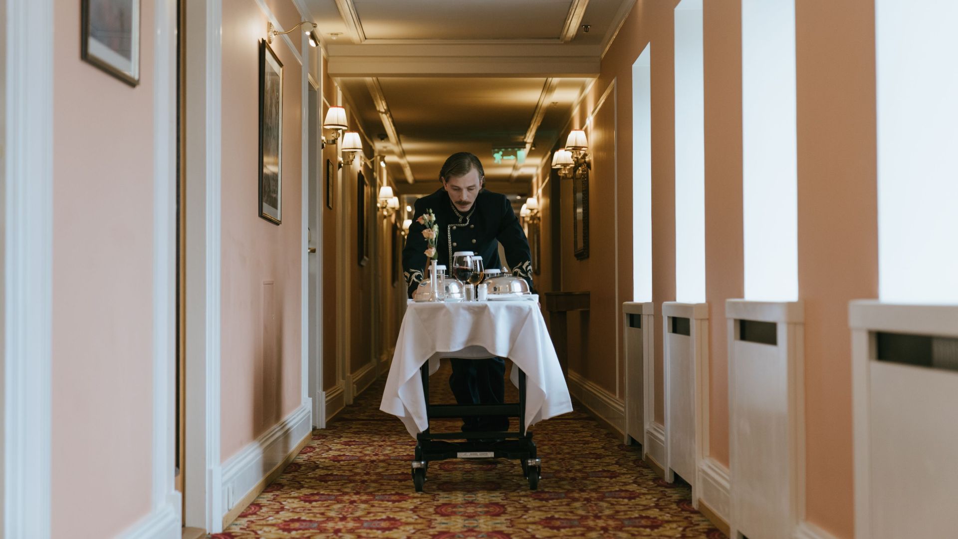 A hotel staff member pushes a room service trolley in a luxurious corridor.