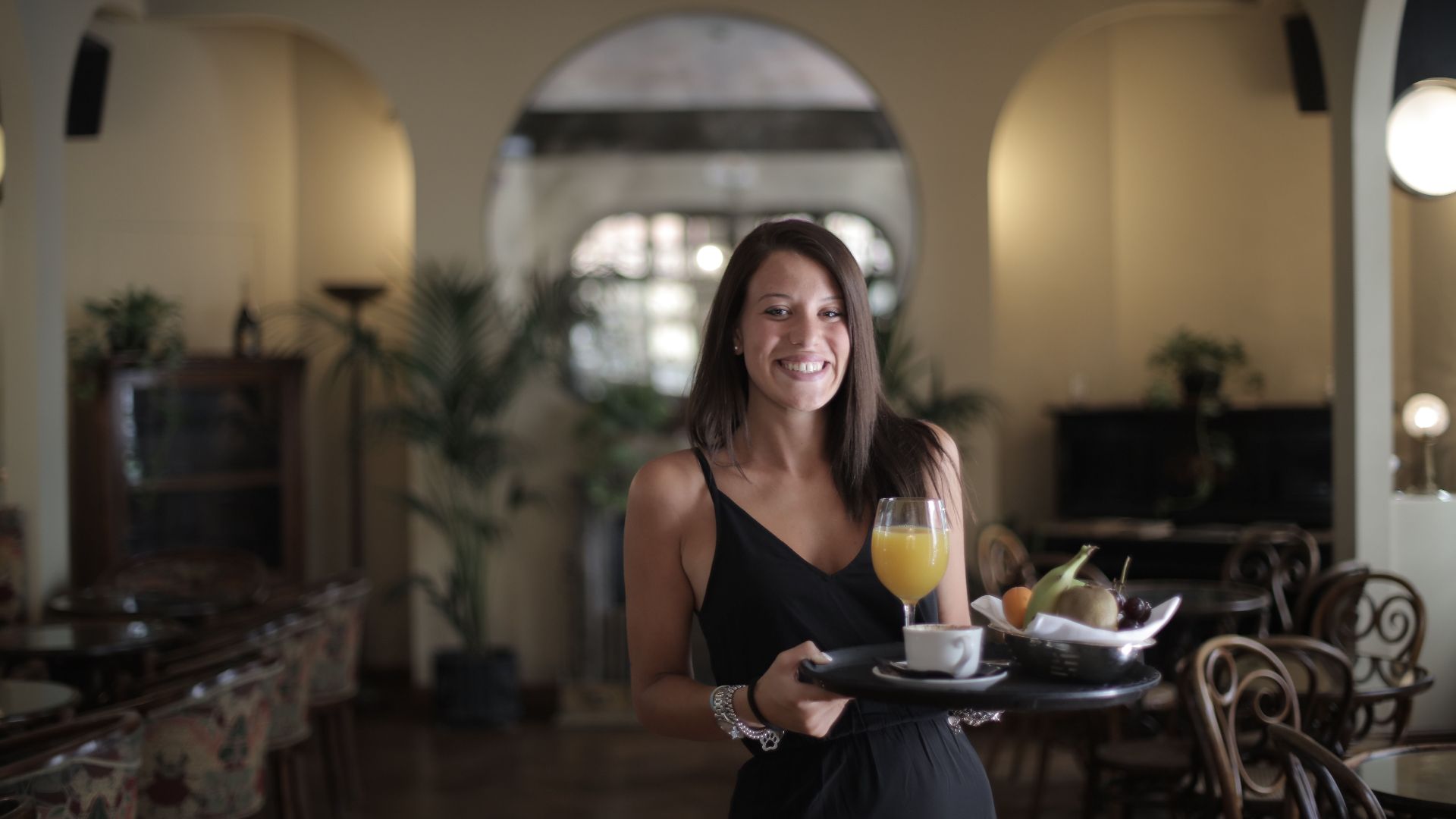 Woman in black dress serving breakfast with juice and fruit in a stylish restaurant.