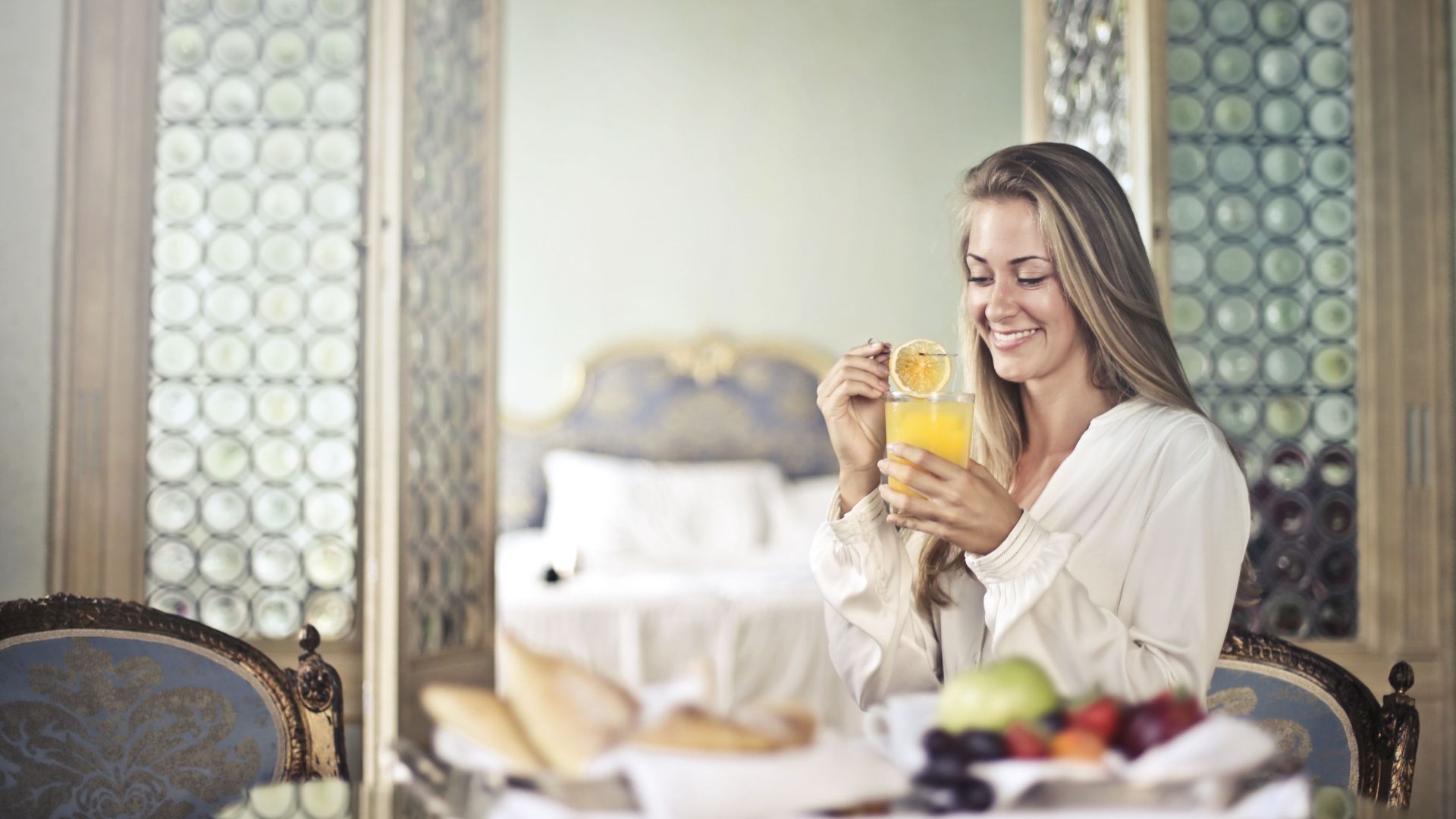 Side view of happy young female sitting at table with glass of fresh juice and enjoying delicious healthy breakfast in cozy room of hotel