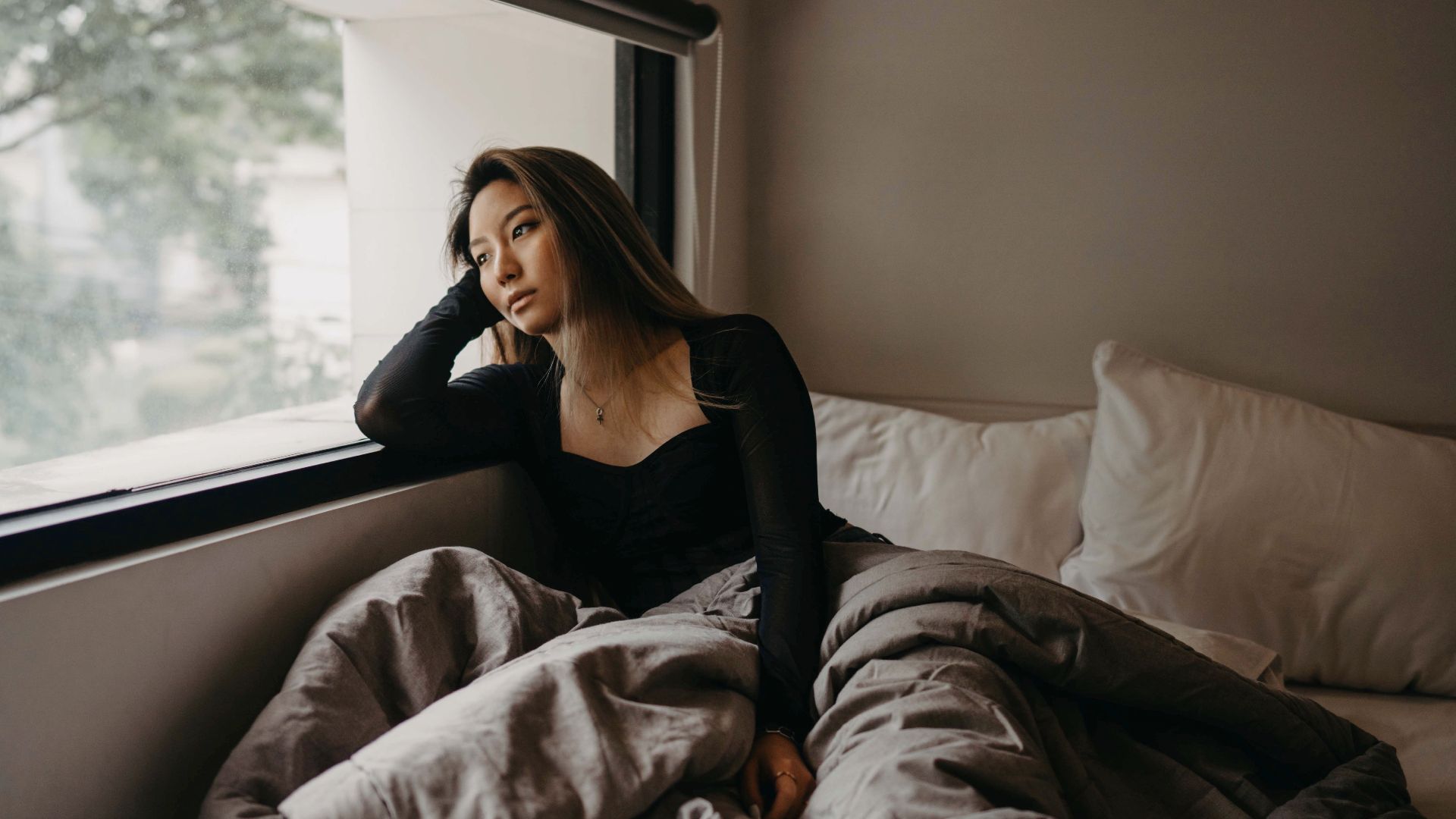 A young woman in a cozy room, pondering by the window, wrapped in blankets.
