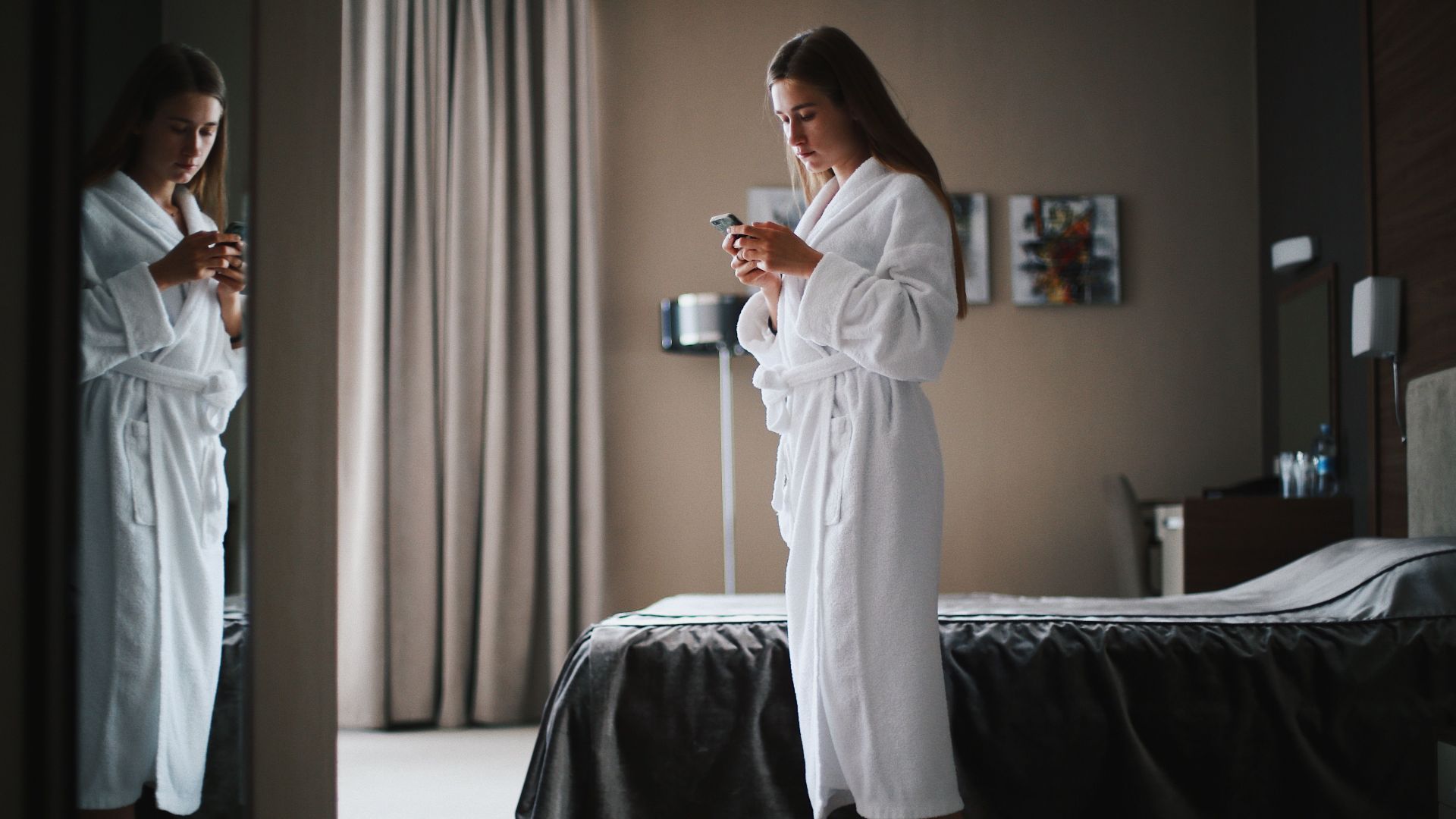 Woman in bathrobe looking at smartphone in a modern hotel room, near a mirror.