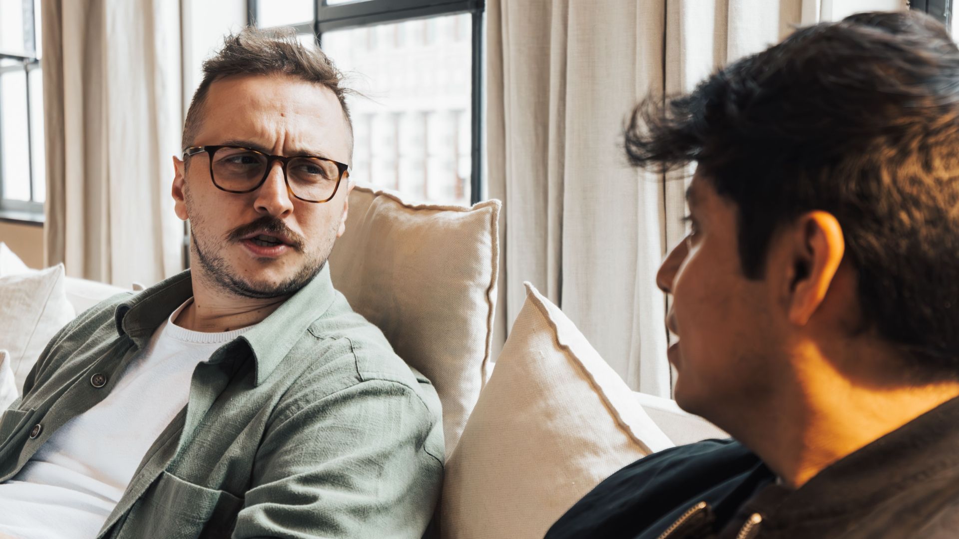 Two men sitting on a sofa, engaging in a conversation while enjoying a coffee indoors.
