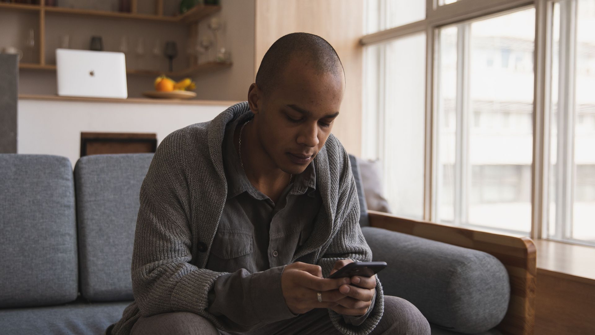 Concentrated young African American man in casual wear relaxing on sofa and surfing internet on mobile phone while sitting near window in cozy apartment