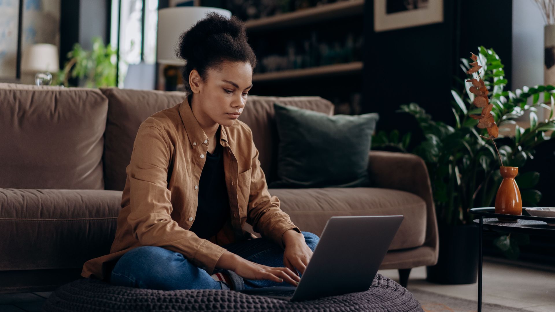 A young woman sitting on a floor cushion using a laptop for remote work in a cozy living room.
