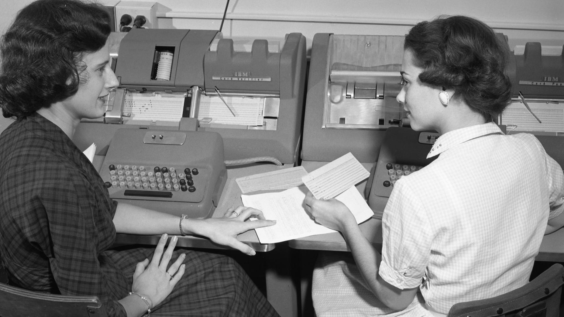 Two women discussing their work while entering data onto punched cards at Texas A&M in the 1950s. The woman at the right is holding a punched card and is seated at an w:IBM 026 keypunch machine.  The woman at left is at an IBM 056 Card Verifier. Her job w