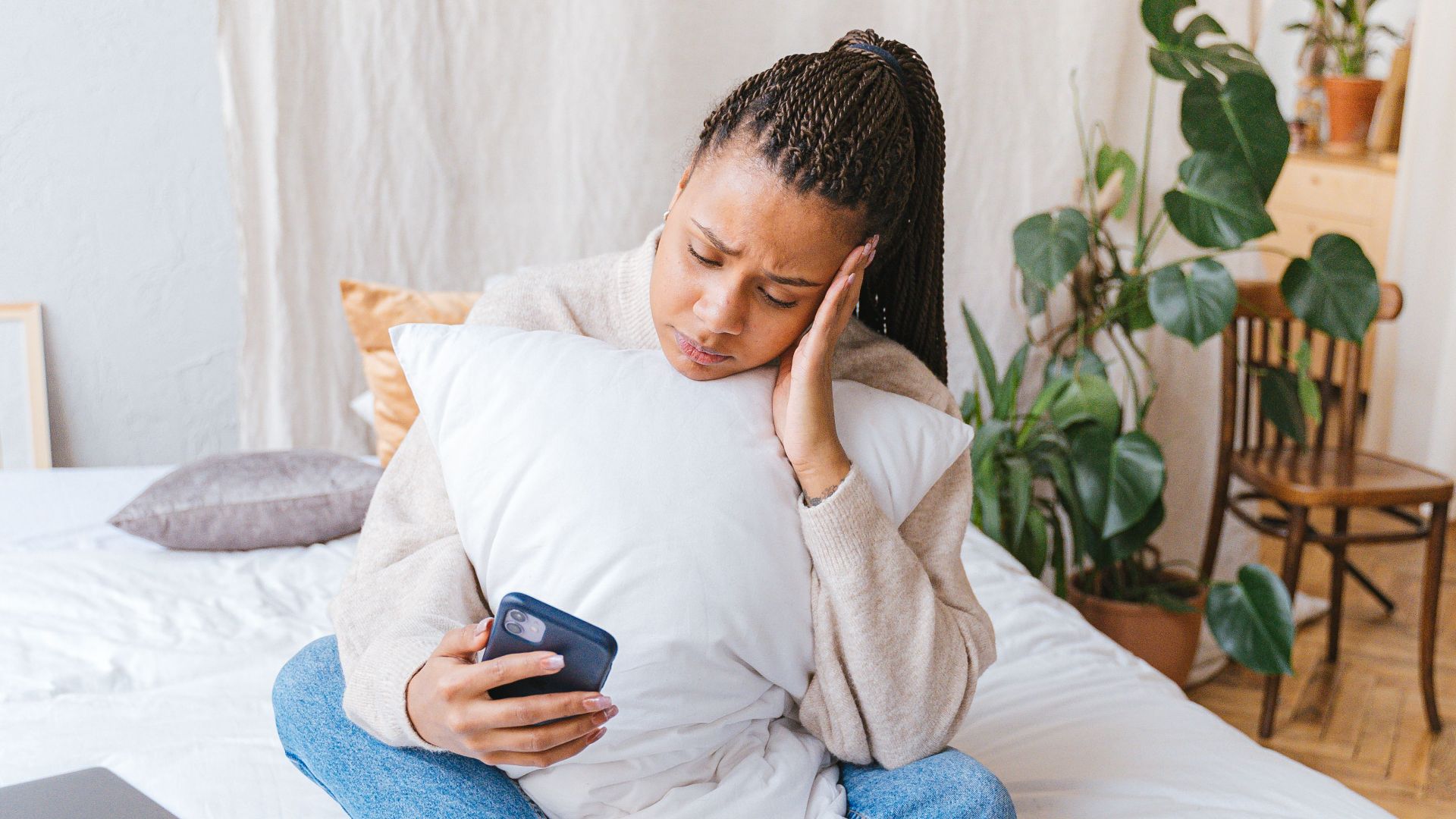Woman sitting on bed, holding smartphone, looking thoughtful. Modern home setting.