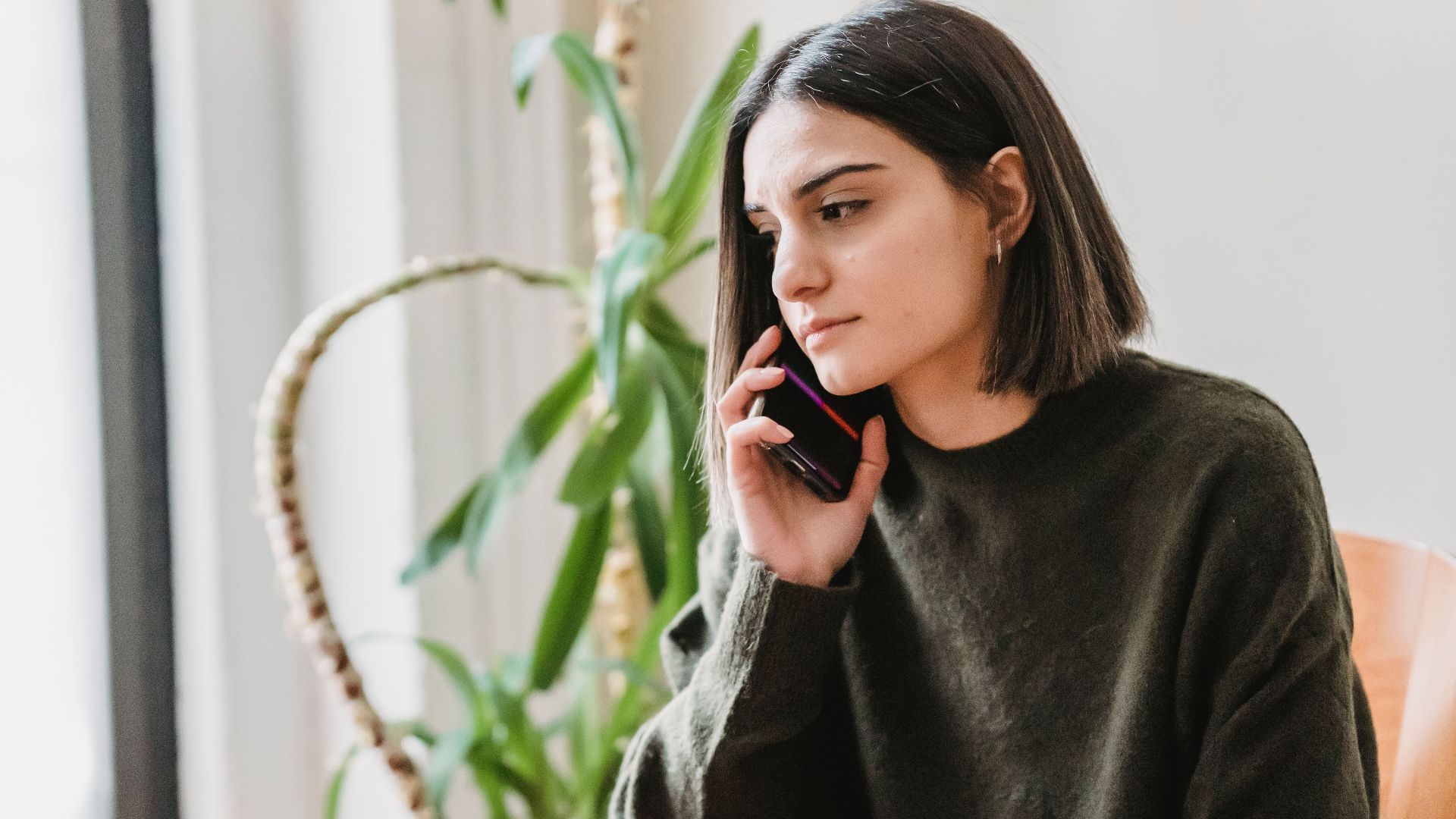 Calm young ethnic female millennial with dark hair in casual clothes having phone conversation while sitting on comfortable armchair at home in daylight