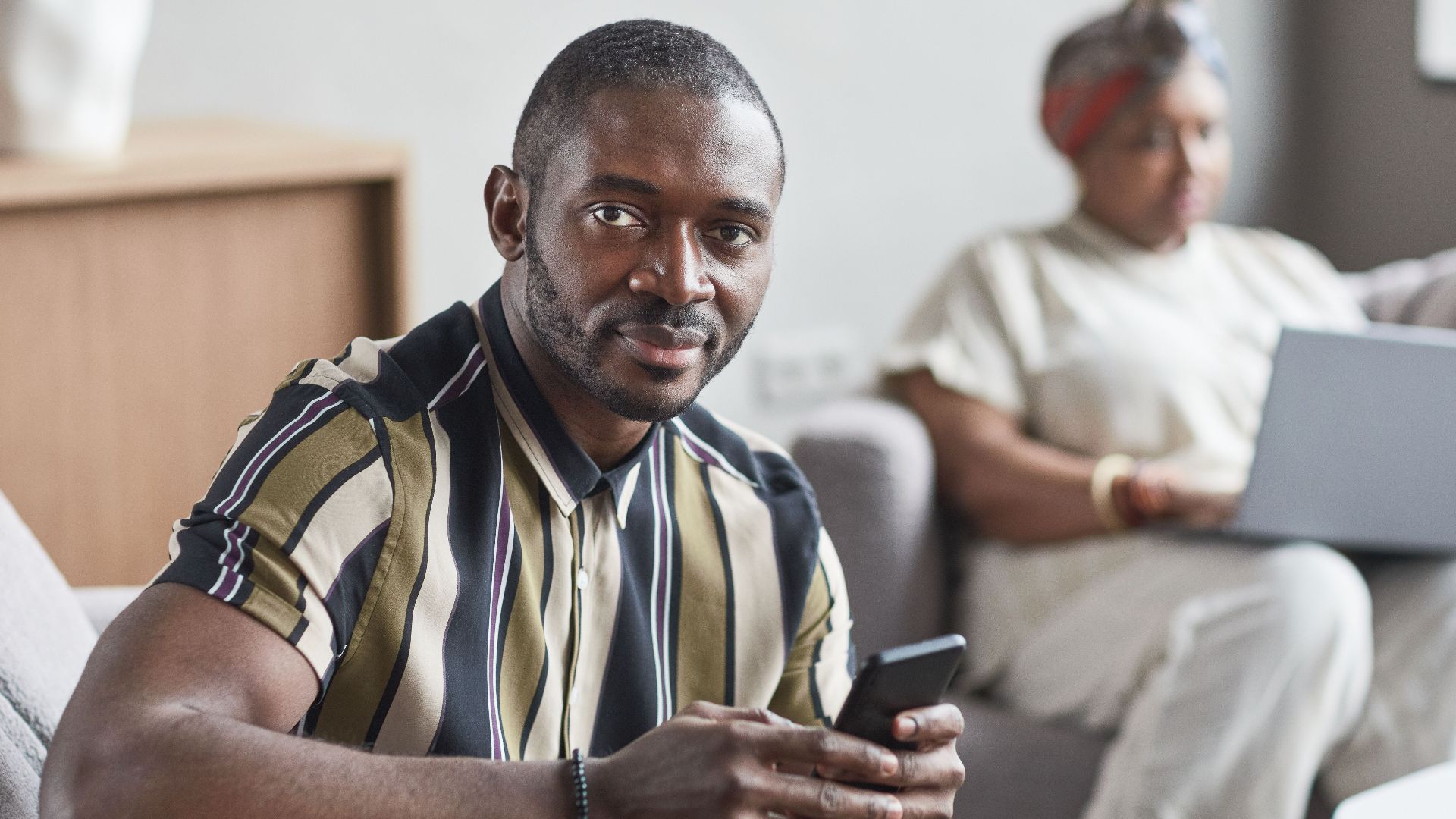 A confident African American man holding a smartphone while sitting indoors.