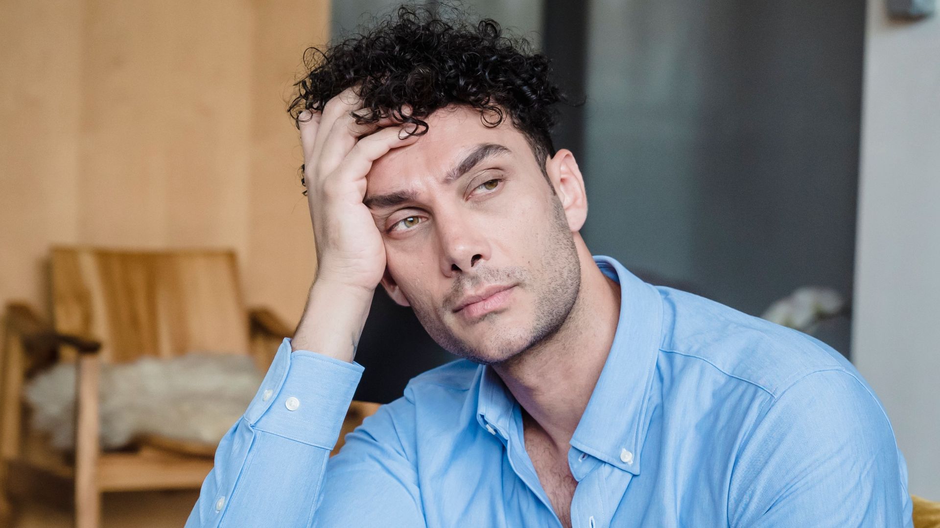 A tired man in a blue shirt listens while a woman talks indoors. Modern living room.
