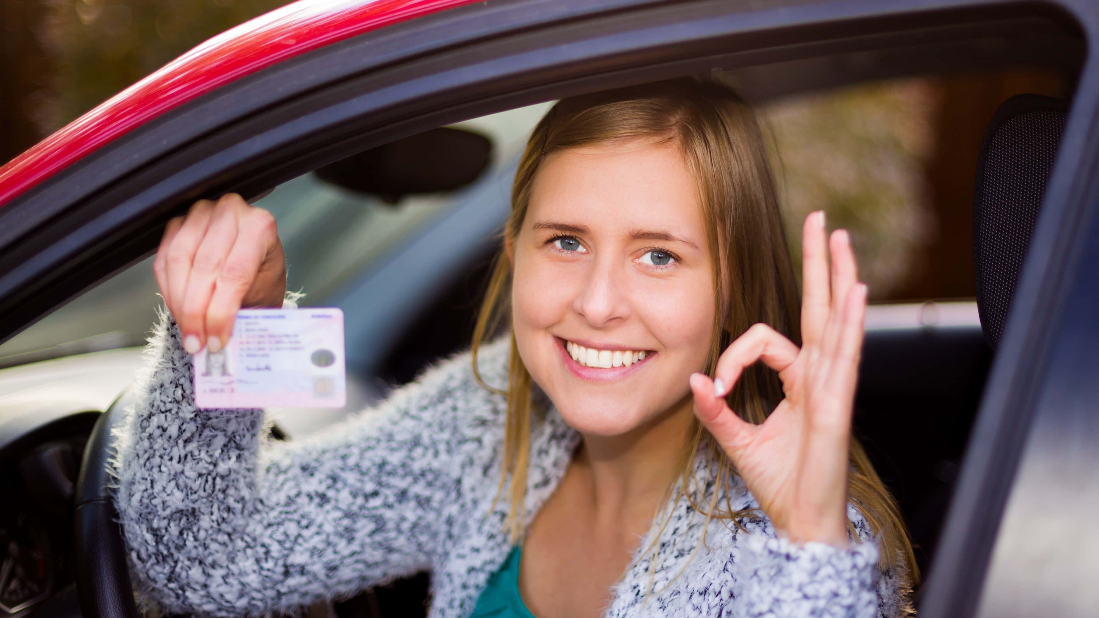 Smiling woman holding her driver license after successful driver's exam in her red car.