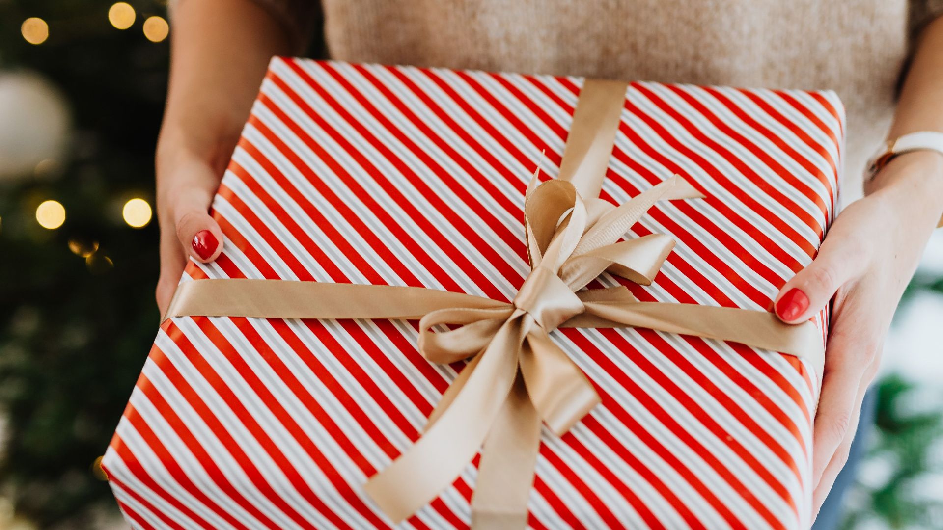A close-up of a person holding a festive wrapped gift with red and white stripes near a decorated Christmas tree.
